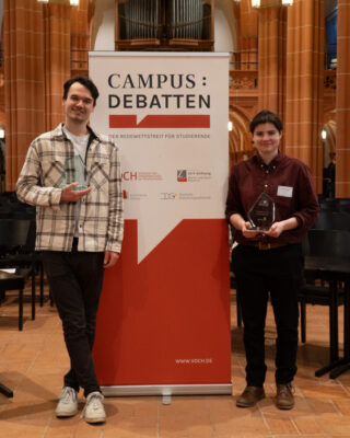 Felix und Agata mit ihren Glaspokalen vor dem Campus-Debatten Banner